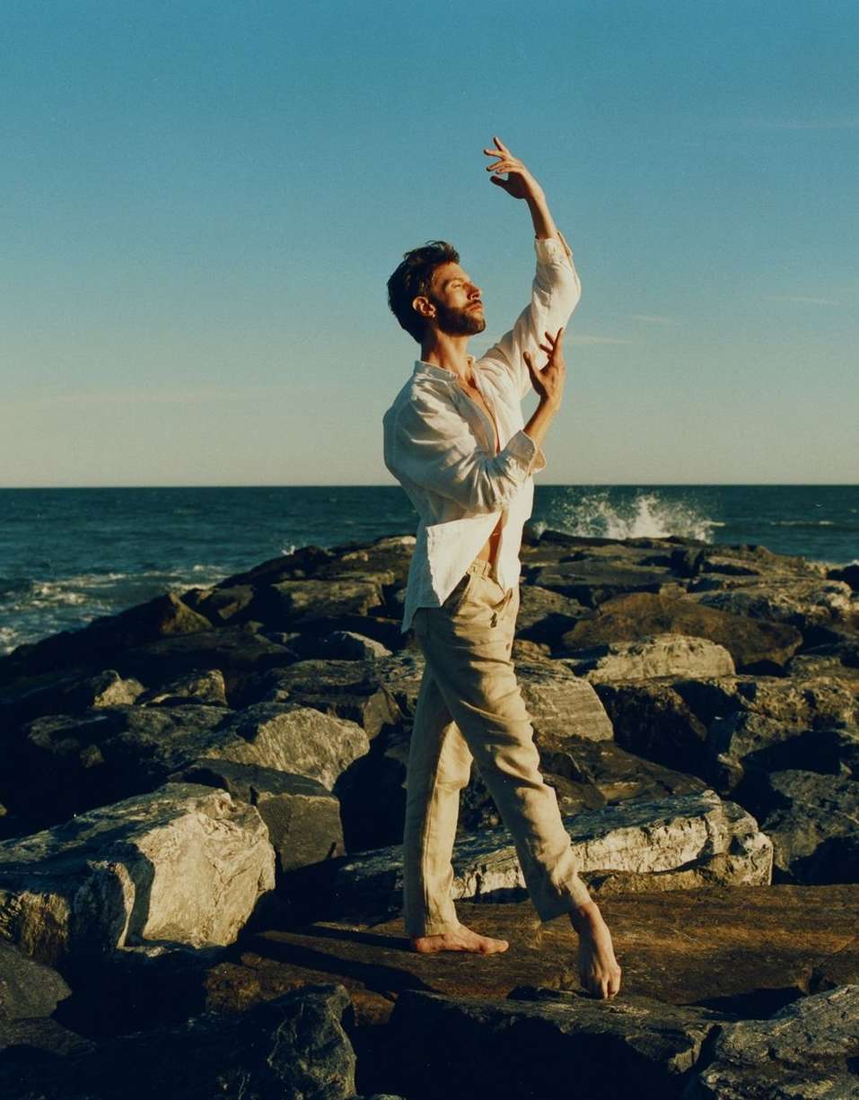 Bearded man in white shirt poses barefoot on rocky pier with arms raised against the ocean and blue sky in outdoor setting.