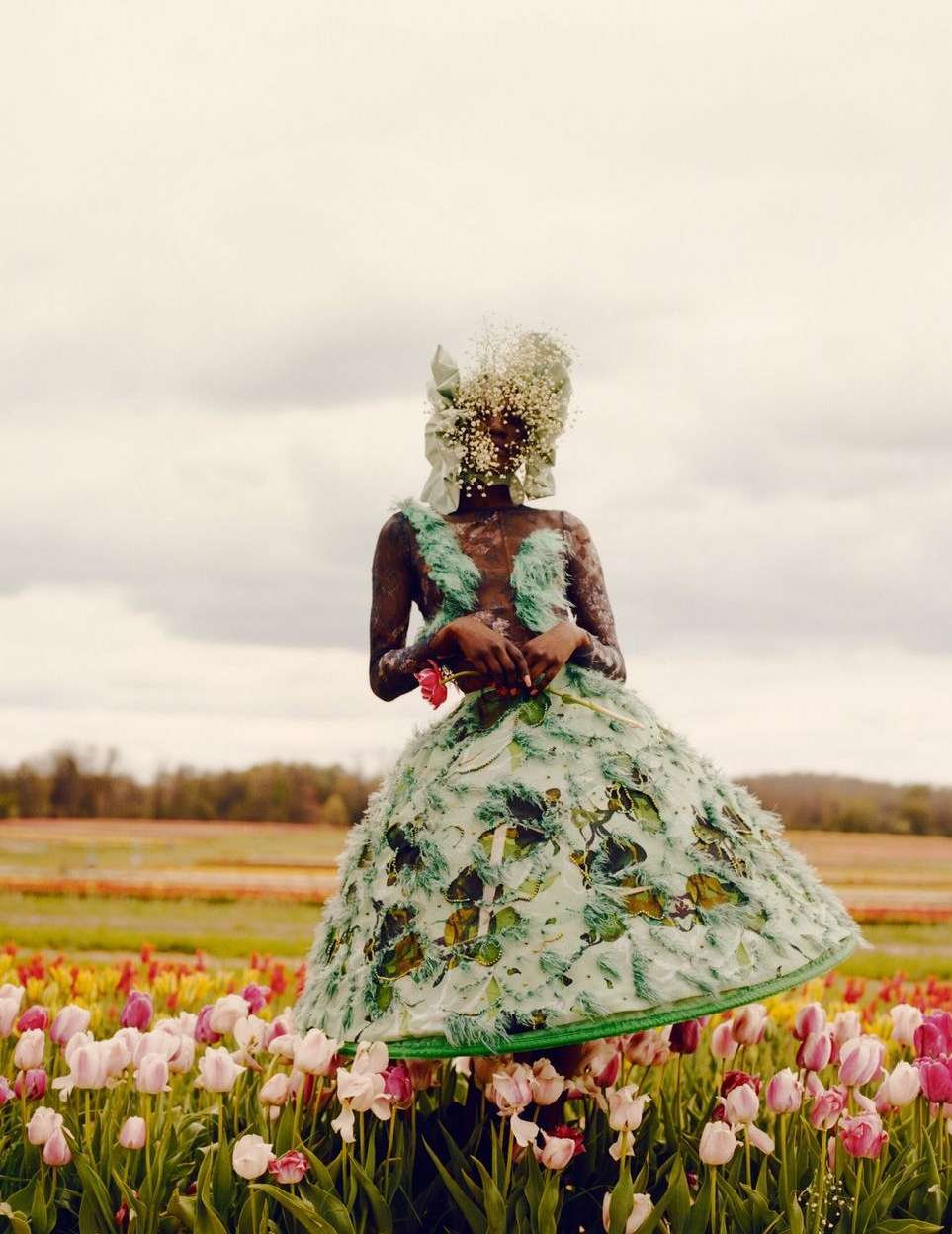 Dark-skinned model in a floral dress stands in a tulip field beneath a cloudy sky, holding a red rose, fashion and floral concept.