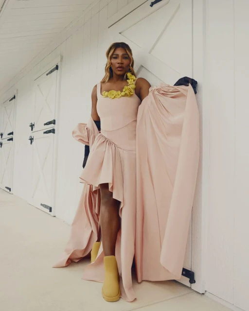 Serena Williams poses in a pink dress with yellow flower detail and tan boots in front of a white paneled wall with closed doors.