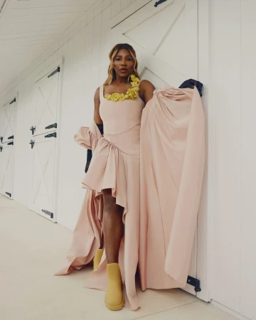 Serena Williams poses in a pink dress with yellow flower detail and tan boots in front of a white paneled wall with closed doors.