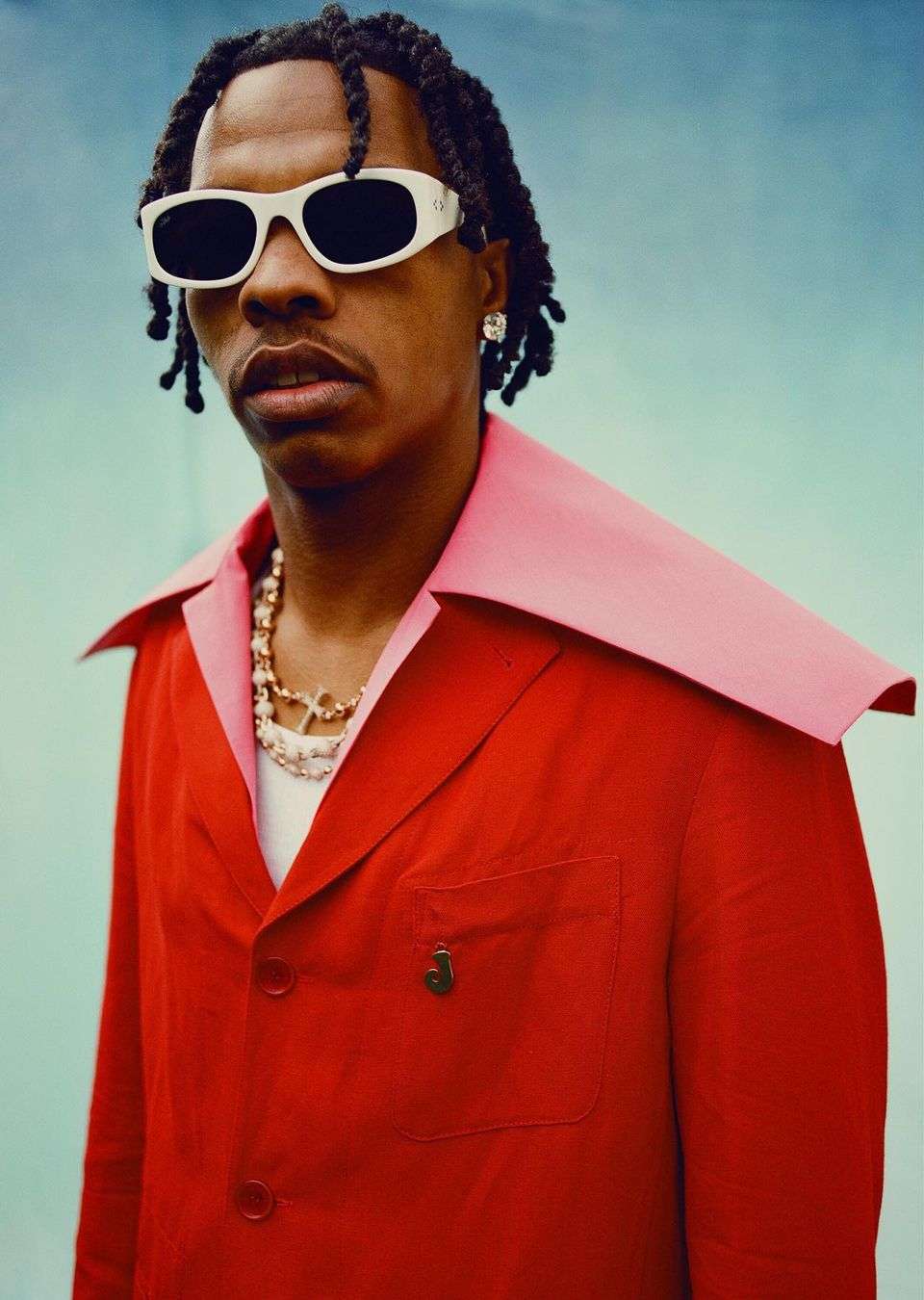 Portrait of Lil Baby in a red suit jacket, white sunglasses, and jewelry, posing in front of a blue backdrop. Medium shot.