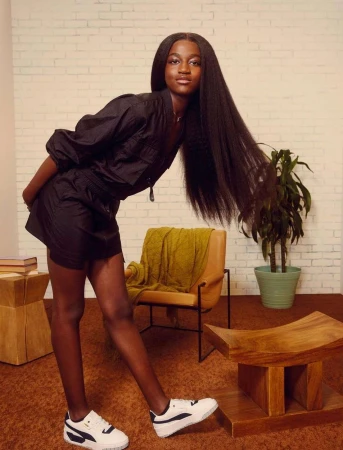 Black woman with long straight hair wearing black outfit and white sneakers posing in a retro interior.