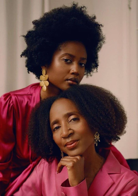 Two women with dark hair and pink jackets pose indoors wearing statement earrings and natural makeup in a fashion portrait.