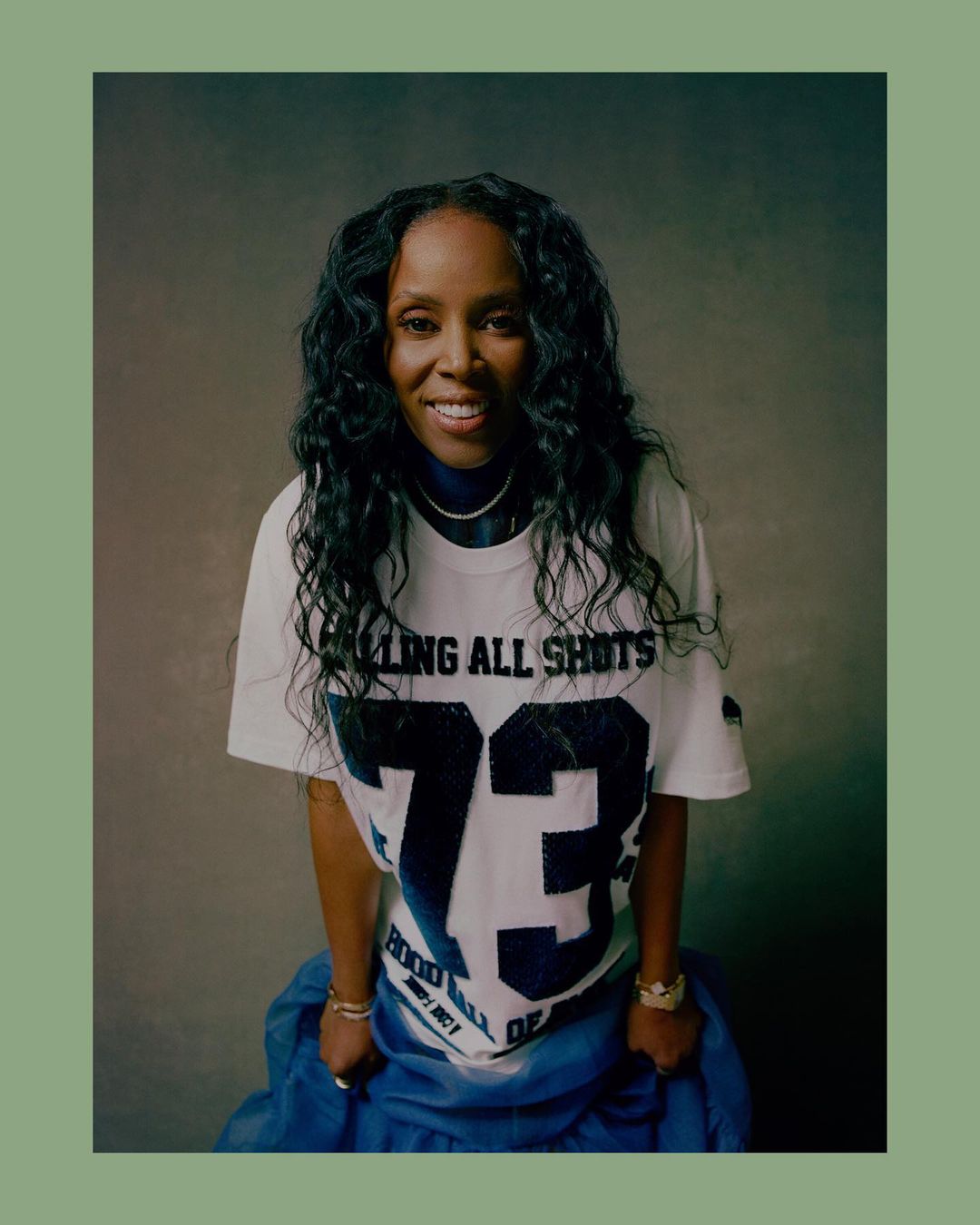 Portrait of a smiling black woman with long dark hair wearing a white graphic t-shirt and blue skirt against a textured grey studio background.