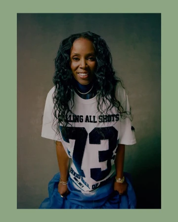 Portrait of a smiling black woman with long dark hair wearing a white graphic t-shirt and blue skirt against a textured grey studio background.
