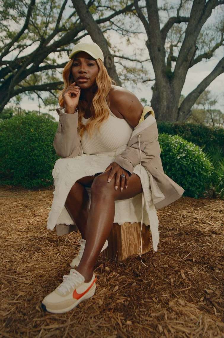 Serena Williams poses in a dress and sneakers, seated on a tree stump in a park-like setting with mature trees and dappled lighting.