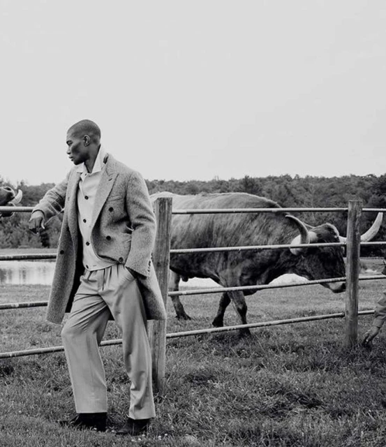 An adult male model in an overcoat leans on a wooden fence with a bull in the background, in a black and white fashion editorial.