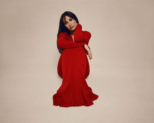 A woman with long dark hair, wearing a long-sleeved red dress, sits on the floor with her hands near her face in a studio setting.