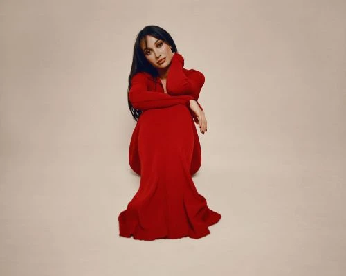A woman with long dark hair, wearing a long-sleeved red dress, sits on the floor with her hands near her face in a studio setting.
