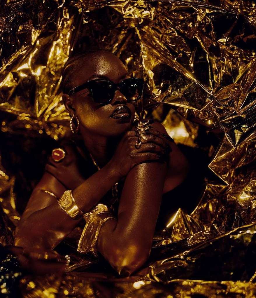 Portrait of a Black woman with sunglasses and gold jewelry, surrounded by gold foil, creating a glamorous, high-contrast fashion image.