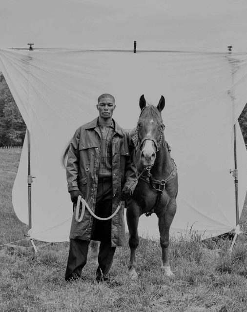 Monochrome portrait of an African American man in a jacket standing with a horse against a backdrop in a grassy field.