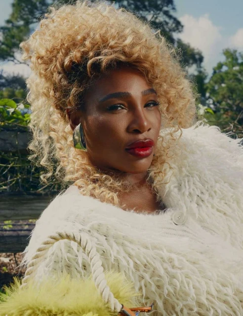 Portrait of Serena Williams with curly blonde hair wearing a white fur-like jacket, posed outdoors with greenery, captured in medium shot.