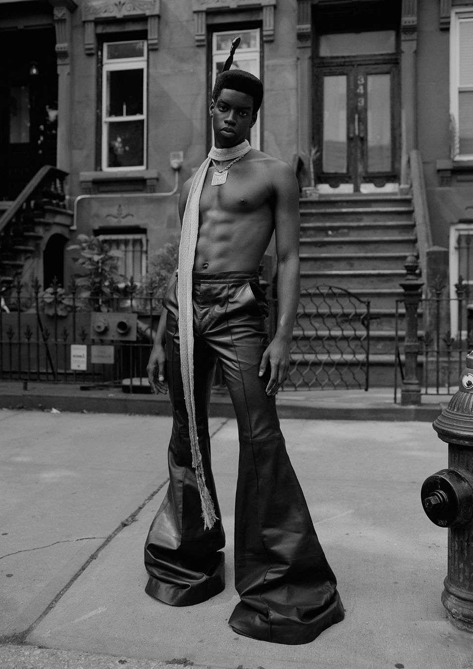 Striking black and white fashion portrait of a shirtless African American man in bell-bottom pants on a city street near brownstones.