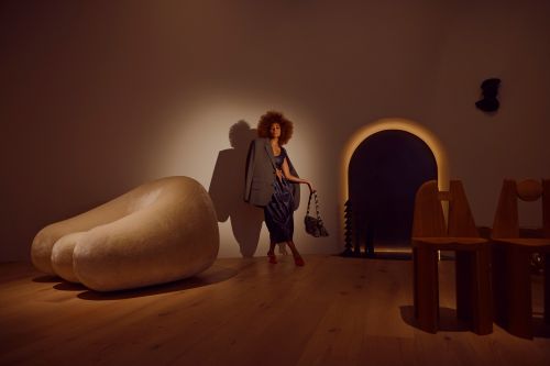 A woman with an afro hairstyle wearing an oversized grey blazer and blue dress poses in a warmly lit gallery interior.