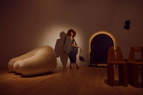 A woman with an afro hairstyle wearing an oversized grey blazer and blue dress poses in a warmly lit gallery interior.