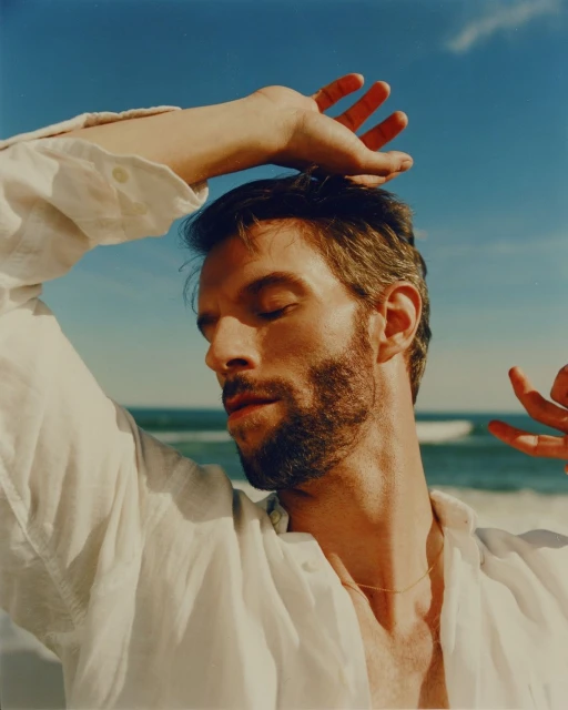 Man with closed eyes poses on the beach with hands above his head. He wears a white linen shirt; blue sky and ocean waves visible.