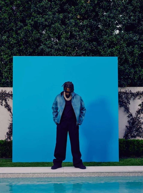 Portrait of a man in a blue jacket and dark pants posing in front of a blue background near a pool with green foliage.