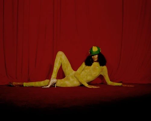 A person lies on their side on a red carpet, wearing a yellow catsuit, a hat, and heels, posing in front of a red curtain backdrop.