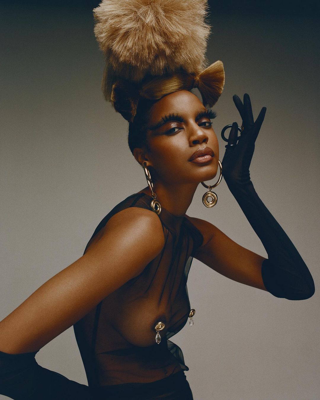 Fashion-forward portrait of a Black woman with a unique updo, dramatic makeup, and large hoop earrings against a grey backdrop.