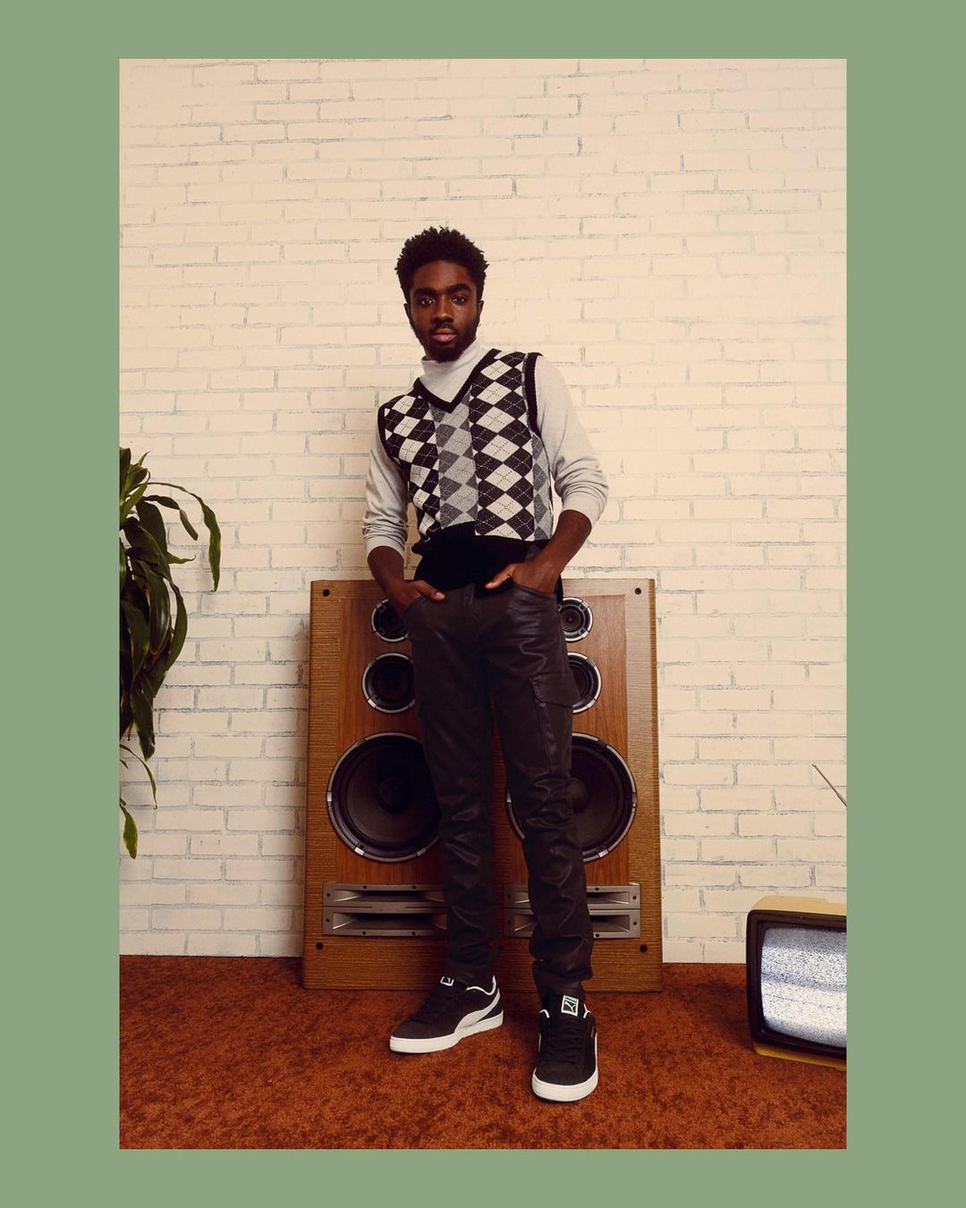A young Black man poses in a gray turtleneck and argyle sweater vest in front of a speaker and brick wall, casual yet stylish.