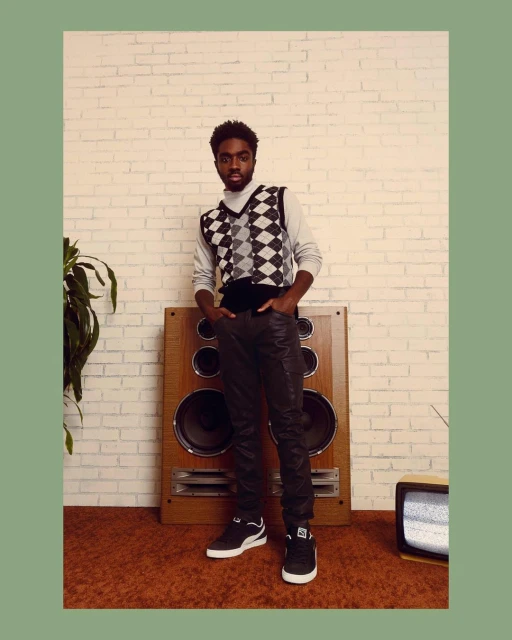 A young Black man poses in a gray turtleneck and argyle sweater vest in front of a speaker and brick wall, casual yet stylish.