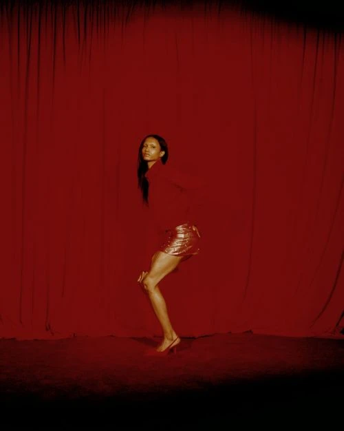 Woman in red top and gold skirt poses in front of draped red fabric in a studio setting with low key lighting and medium shot.