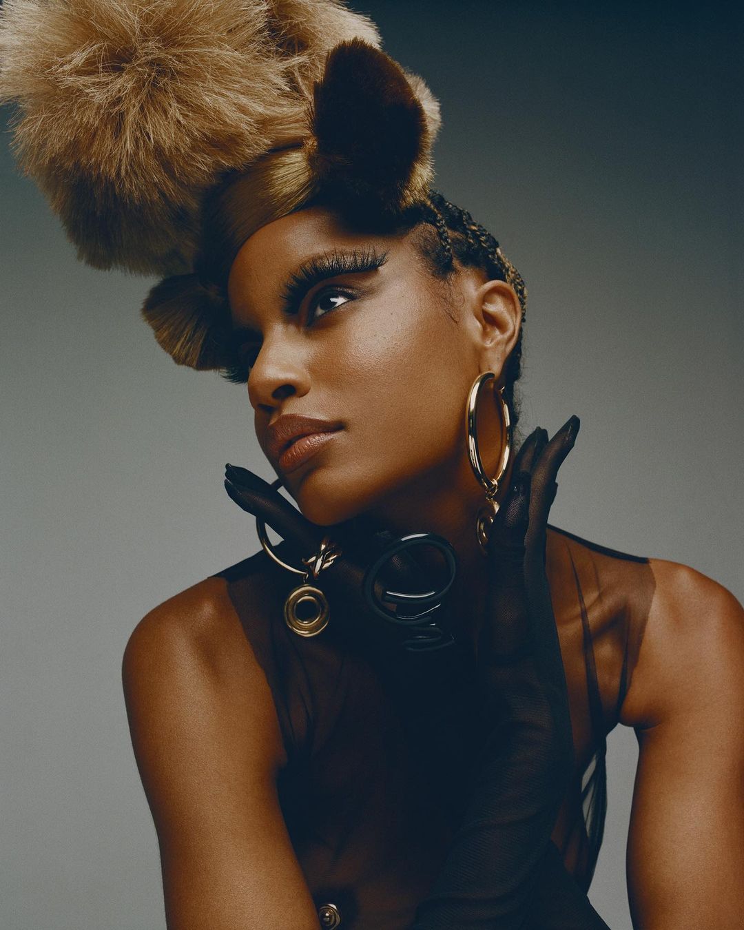 Portrait of a stylish African-American woman with braids and dramatic eyelashes wearing hoop earrings, sheer top, and fuzzy headpiece.