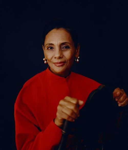 Portrait of a woman in a red jacket sitting with hands clasped on a chair against a dark background. The woman is smiling slightly.