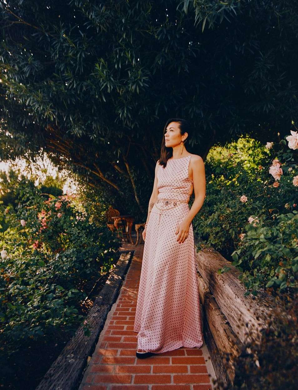 Woman in polka dot dress stands on brick path surrounded by lush greenery and blooming flowers, looking off into the distance.