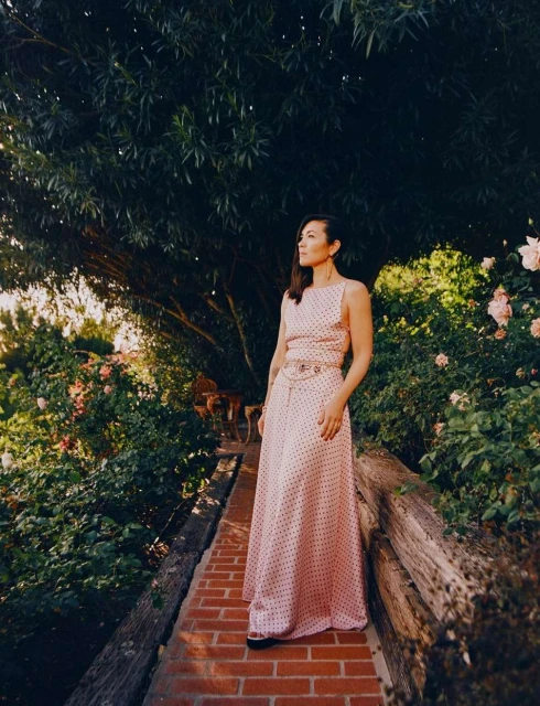 Woman in polka dot dress stands on brick path surrounded by lush greenery and blooming flowers, looking off into the distance.
