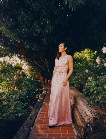 Woman in polka dot dress stands on brick path surrounded by lush greenery and blooming flowers, looking off into the distance.