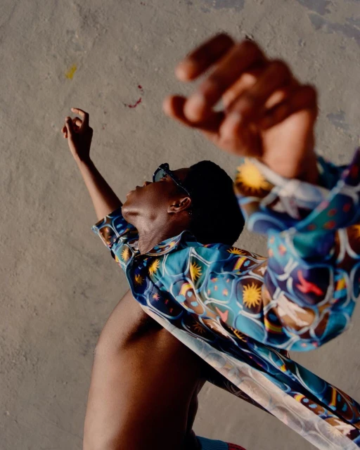 Adult with raised arms wears a patterned shirt, sunglasses. The background is a textured wall in warm sunlight. Fashion editorial.