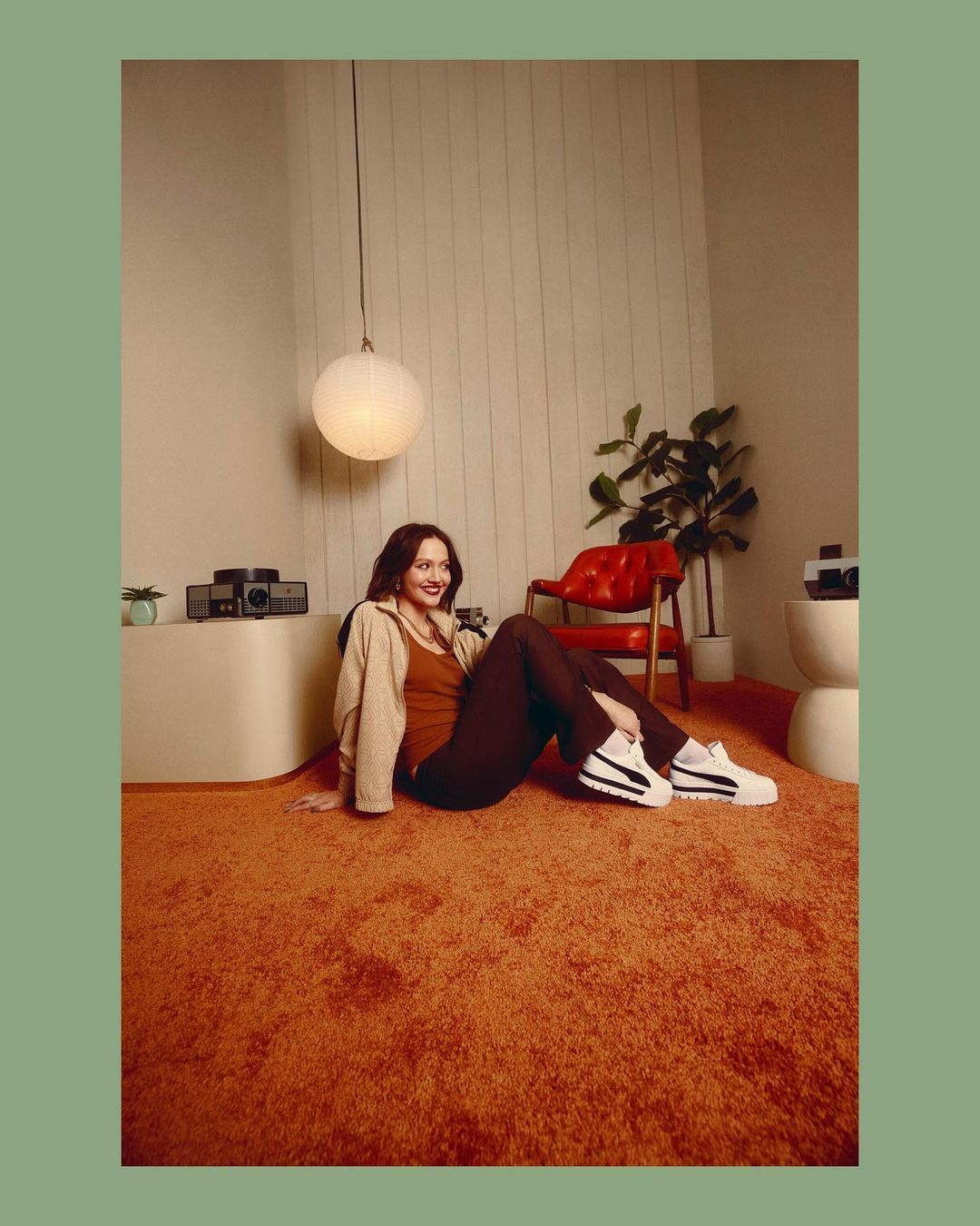 A woman in a brown tank top and pants, wearing Puma sneakers, sits on an orange shag rug, with mid-century modern decor in the background.