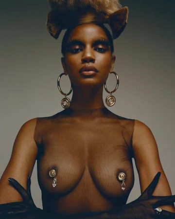 Close-up studio shot of a dark-skinned woman with creative makeup, gold hoop earrings, and hand jewelry against a gray background.