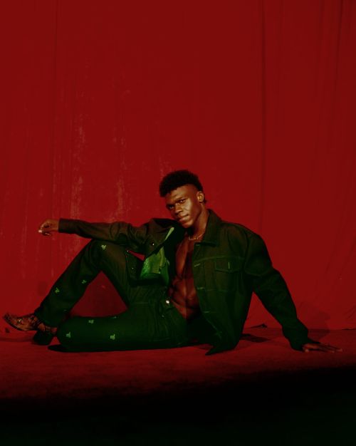Black man in a green jacket and trousers seated against a textured red background