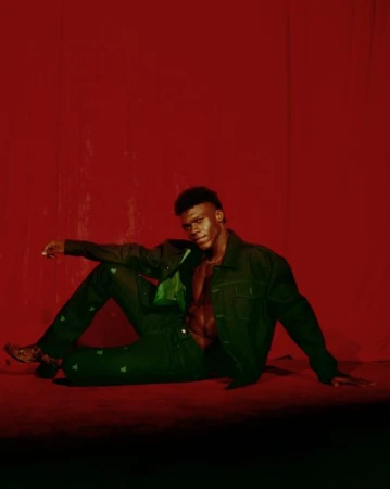 Black man in a green jacket and trousers seated against a textured red background