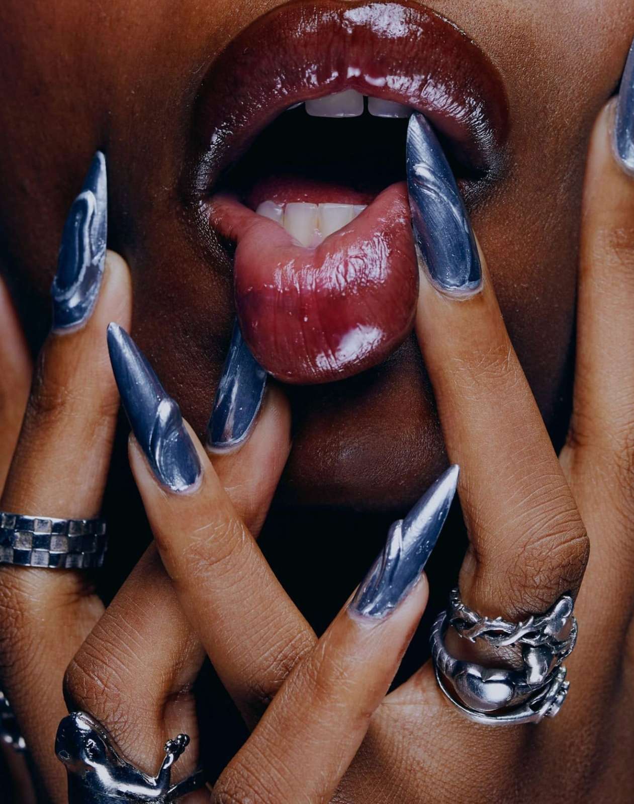 Macro shot of a person with dark skin and glossy lips displaying an extended tongue and silver nail polish with chrome rings.