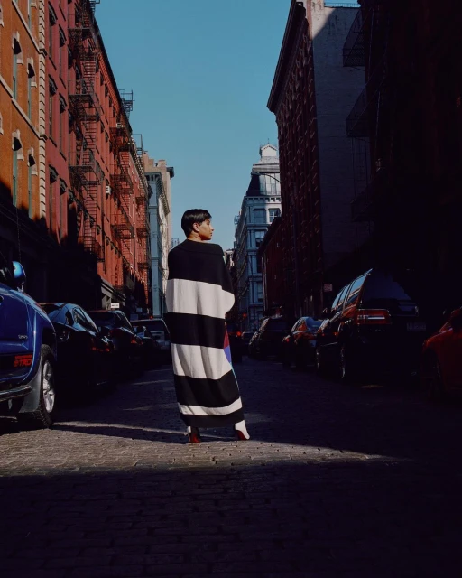 A woman in a striped robe stands on a city street lined with parked cars and brick buildings under a clear blue sky in urban setting.