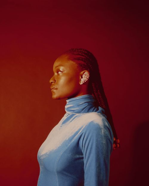 Portrait of a stylish Black woman in a blue velvet turtleneck and ornate earring, standing in front of a red background, facing left.