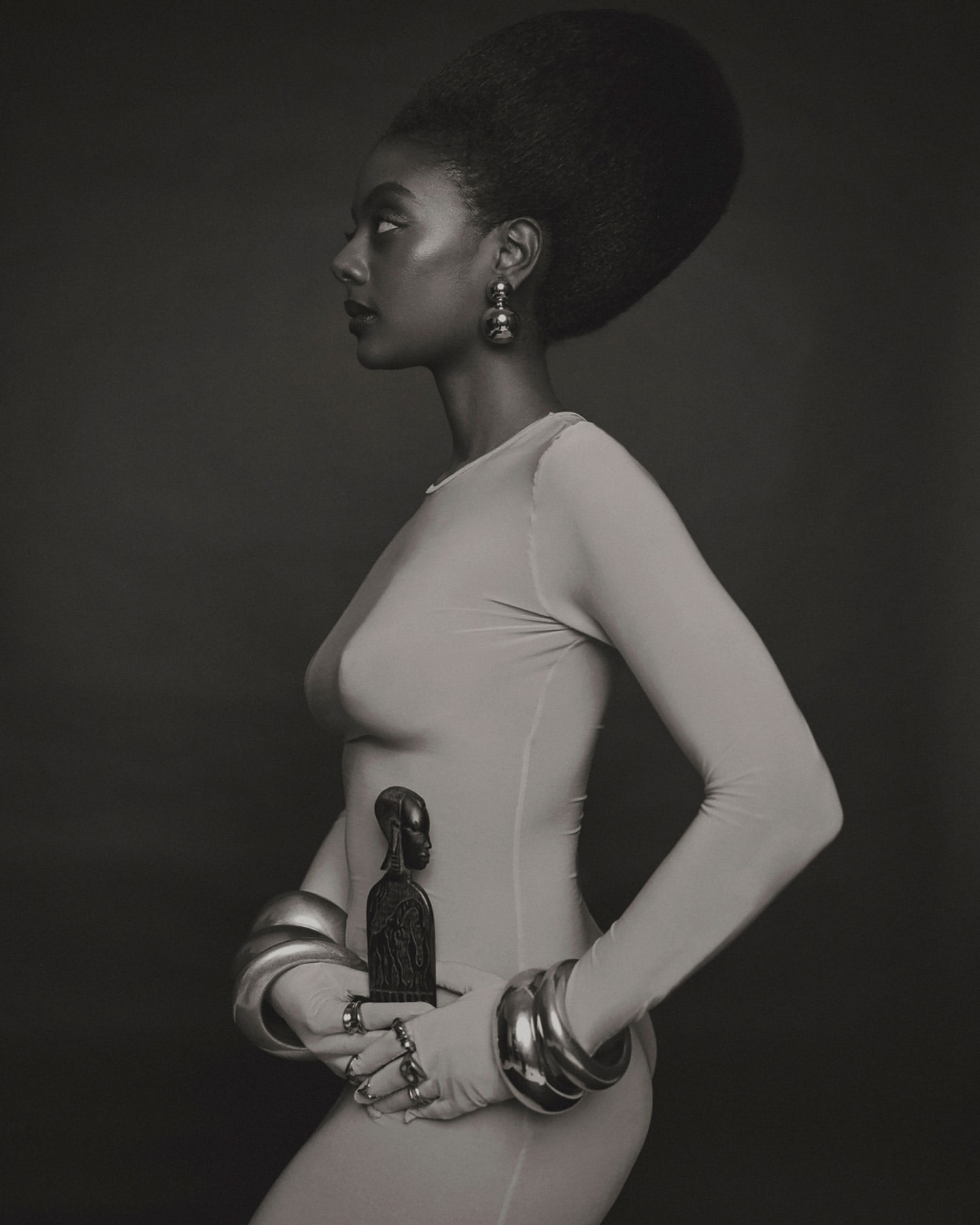 Profile portrait of an African American woman with an afro in a light dress, wearing several bangles and holding a dark statue.