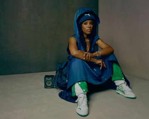Adult female in a blue hooded outfit and white sneakers sitting indoors next to a vintage boombox and backpack against a neutral wall.