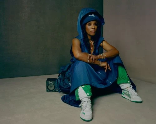 Adult female in a blue hooded outfit and white sneakers sitting indoors next to a vintage boombox and backpack against a neutral wall.