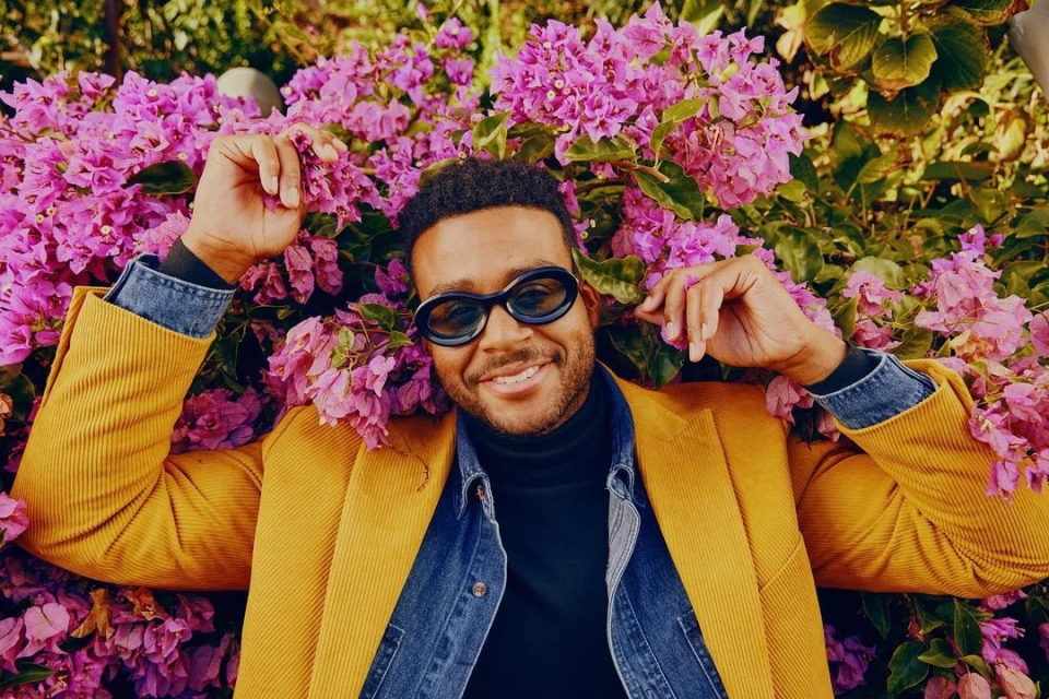 Smiling man in sunglasses and yellow jacket surrounded by pink bougainvillea flowers. Outdoor fashion portrait in natural light.