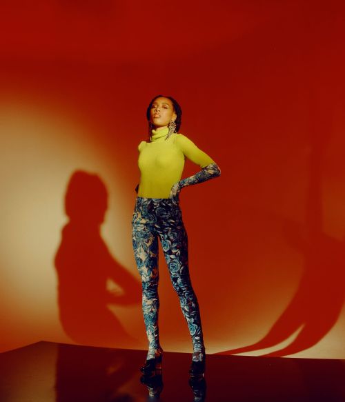Fashion shot of an African-descent woman in a yellow top and patterned pants, standing in a photo studio with orange background lighting.