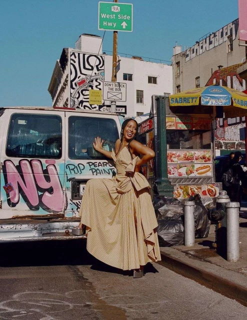 A woman in a dress poses next to a graffiti-covered van with a food stand in the background, creating a vibrant street fashion scene.