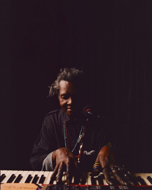 African-American man plays electronic piano in dark lighting, showcasing hands, keyboard, microphone, and dark background.