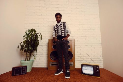 Full-length portrait of a Black man in patterned sweater vest standing indoors next to vintage TV and speaker on shag carpet.