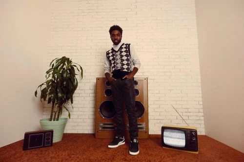 Full-length portrait of a Black man in patterned sweater vest standing indoors next to vintage TV and speaker on shag carpet.