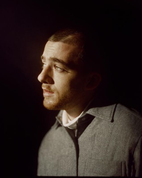 Portrait of a young adult man with short brown hair, gazing slightly upward in a dimly lit, dark environment. He is wearing a collared jacket.