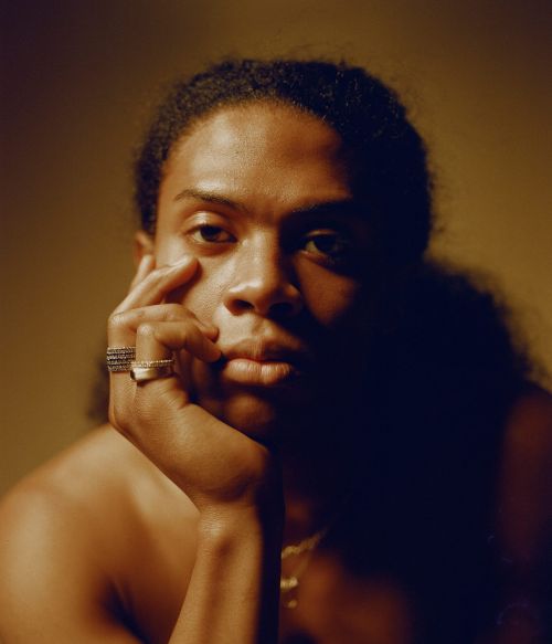 Portrait of a black man resting his chin on his hand with rings on his fingers against a warm-toned background
